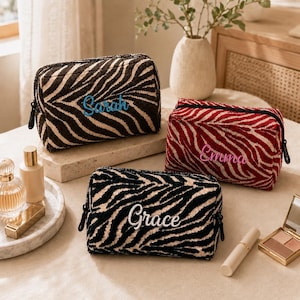 Op de afbeelding: Drie rechthoekige make-uptasjes met een gestructureerd zebrastreepmotief. Eén tas is bruin en beige met de naam Sarah in het blauw, één is rood en beige met de naam Emma in het roze, en één is zwart en beige met de naam Grace in het wit.