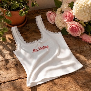 Op de afbeelding: Een wit geribbelde tanktop met een ronde halslijn, afgewerkt met delicate witte kanten randen en een klein strikje in het midden. De tekst "Mrs. Destiny" is in rode schrijfletters op de voorzijde van het kledingstuk geborduurd in ribstof.