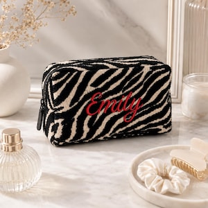 May include: A rectangular cosmetic bag featuring a black and white zebra print pattern. The front of the bag is embroidered with the name Emily in a red cursive script. It includes a black zip closure with a matching black fabric pull tab.