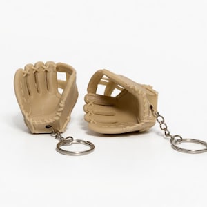Puede incluir: Dos llaveros de guante de béisbol color beige. Cada guante en miniatura tiene costuras detalladas y una cadena de metal con un llavero. Los guantes están colocados sobre un fondo blanco.