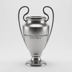 Puede incluir: Una copa de trofeo plateada con dos asas curvas. La copa tiene un cuerpo ancho y redondeado y una base de pedestal. Las palabras "COUPE DES CLUBS CHAMPIONS EUROPEENS" están grabadas en la copa. El trofeo está sobre un fondo blanco.