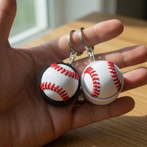 May include: Two baseball-themed keychains. Each keychain features a miniature baseball with red stitching on a white surface. One baseball is set in a black base, the other in a white base. Both have silver keyrings.