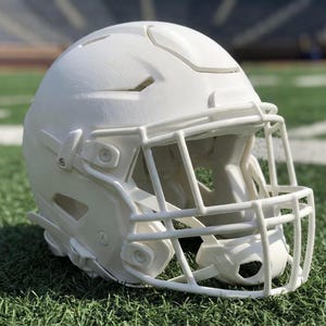 Könnte beinhalten: Ein weißer American-Football-Helm mit Gesichtsmaske, der auf einem grünen Rasenfeld liegt. Der Helm hat eine glatte, saubere Oberfläche mit Belüftungsöffnungen und einem stabilen Gesichtsschutz. Der Hintergrund zeigt ein Stadion.