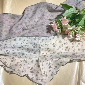 May include: Two rectangular scarves featuring a delicate floral pattern in shades of gray and white. The fabric has a textured appearance with dark gray scalloped edges. A cluster of artificial pink and white flowers rests on the scarves.