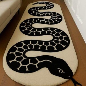 Peut inclure: Un tapis ovale allongé présentant un fond crème avec un grand motif de serpent noir sinueux. Le serpent possède une tête noire distincte avec une langue bifide et un corps orné d'écailles noires de forme hexagonale sur toute sa longueur.