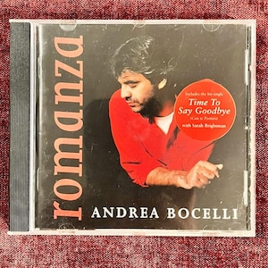 Może przedstawiać: Etui na płytę CD z albumem Romanza Andrei Bocellego. Okładka przedstawia zdjęcie osoby w czerwonej koszuli na czarnym tle. Okrągła czerwona naklejka z przodu zawiera napis: Includes the hit single Time To Say Goodbye.