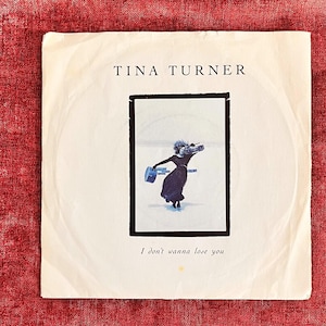 May include: A square vinyl record sleeve featuring the text TINA TURNER at the top and I don't wanna lose you at the bottom. The central image shows a person in a black dress carrying a boombox and a blue case against a light background.