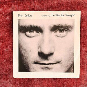 May include: A square vinyl record sleeve featuring a black and white close-up portrait of Phil Collins. The text Phil Collins and In The Air Tonight ('88 Remix) is printed in black ink at the top of the white border surrounding the central image.