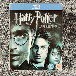 Puede incluir: Una caja de discos Blu-ray titulada Harry Potter The Complete 8-Film Collection. La portada presenta tres retratos del personaje principal con gafas redondas, sobre un fondo verde azulado oscuro con el logo de Blu-ray y dos clasificaciones de edad 12.