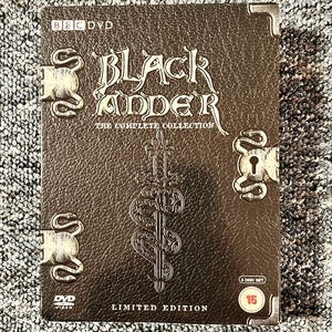 May include: A brown, textured DVD box set titled Blackadder The Complete Collection. The cover features a central sword and snake emblem, metallic corner accents, and side latches. It includes the text BBC DVD, 6 Disc Set, and Limited Edition.