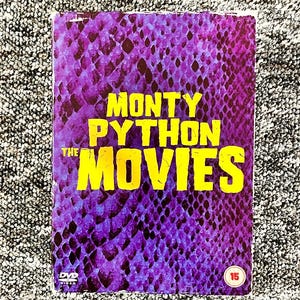 May include: A rectangular DVD case featuring a purple and black snakeskin pattern background. The text MONTY PYTHON THE MOVIES is printed in large, bold yellow letters across the center. A small DVD Video logo and a circular 15 age rating are visible.