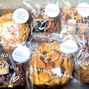 May include: A collection of six individually wrapped gourmet cookies in a box, each featuring distinct toppings like marshmallows, chocolate chunks, and cookie crumbles, with white circular labels and handwritten messages on the clear plastic packaging.