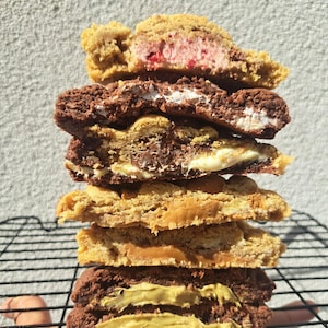 May include: A vertical stack of six thick, halved cookies featuring various fillings. The layers include a pink berry centre, a white marshmallow-like filling, a dark chocolate ganache, a golden caramel, a smooth peanut butter, and a green pistachio cream.