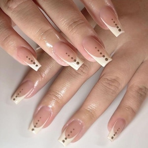 Könnte beinhalten: Lange, quadratisch geformte Fingernägel im klassischen French-Manicure-Stil mit nudefarbener Basis und weißen Spitzen. Jeder Nagel ist mit einer vertikalen Linie aus kleinen, dunkelbraunen Punkten akzentuiert, die der Kurve der weißen Spitze folgen.