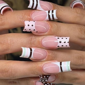 Puede incluir: Uñas postizas largas de forma cuadrada con estilo de manicura francesa sobre una base color nude. Las puntas están decoradas con varios patrones que incluyen lunares marrones, rayas marrones sólidas y bandas horizontales alternas en verde claro y blanco.