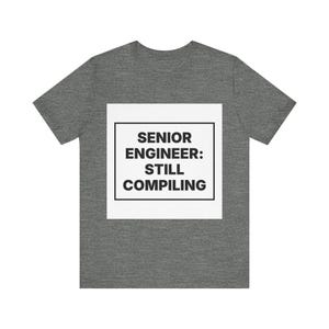 Peut inclure: Un t-shirt à manches courtes gris chiné foncé avec un motif carré blanc sur la poitrine. À l'intérieur du carré, une bordure rectangulaire noire entoure le texte noir en gras qui indique : SENIOR ENGINEER: STILL COMPILING en police sans-serif.
