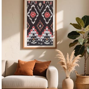 May include: A framed textile art piece with a dark background and a repeating pattern of white, red, and black geometric shapes. The artwork is displayed above a beige sofa with two rust-colored throw pillows. A vase with dried pampas grass and a potted plant are also in the room.