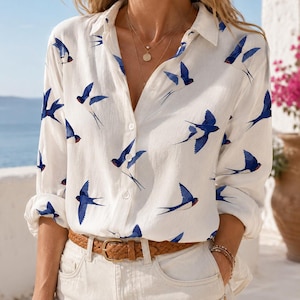 May include: A white long-sleeve button-down shirt featuring a repeating pattern of blue and black swallow birds in flight. The shirt has a classic collar, a front button placket, and is tucked into light-colored pants with a brown braided belt.