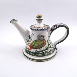 May include: A white ceramic teapot featuring a hand-painted bird with green, orange, and grey plumage. The teapot has a wide base, a curved handle with dark green stripes, and a lid with a pointed finial. Green foliage and blue flowers are painted around the bird, with dark green decorative borders along the base and rim.