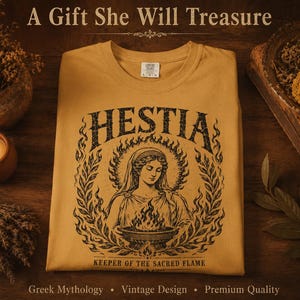 May include: Mustard-colored t-shirt featuring a black graphic of Hestia, the Greek goddess of the hearth, surrounded by flames and laurel leaves. The text "HESTIA" and "KEEPER OF THE SACRED FLAME" are also displayed. The shirt is on a wooden surface.