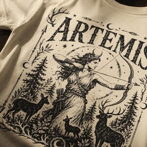 May include: Cream-colored t-shirt featuring a black graphic of Artemis, the Greek goddess of the hunt, with a bow and arrow. The design includes deer, trees, and the word "ARTEMIS" in a distressed font. Arrows and stars complete the design.
