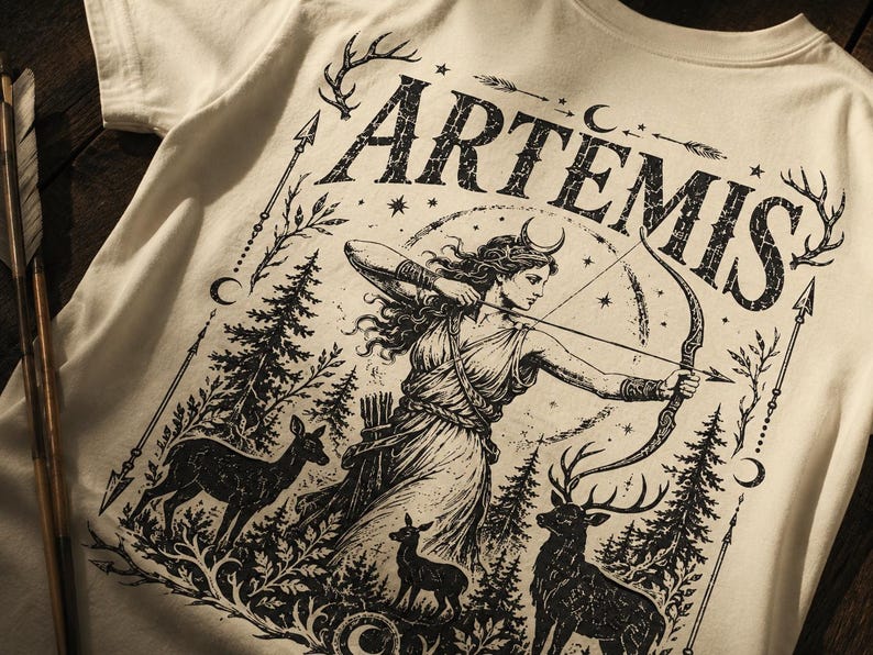 May include: Cream-colored t-shirt featuring a black graphic of Artemis, the Greek goddess of the hunt, with a bow and arrow. The design includes deer, trees, and the word "ARTEMIS" in a distressed font. Arrows and stars complete the design.