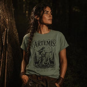 May include: Sage green t-shirt featuring a graphic of Artemis, the Greek goddess of the hunt, with a bow and arrow, deer, and forest elements. The word "ARTEMIS" is at the top of the design. The shirt is worn by a person with dark hair.