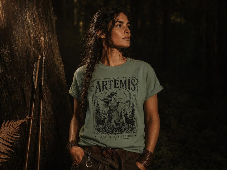 May include: Sage green t-shirt featuring a graphic of Artemis, the Greek goddess of the hunt, with a bow and arrow, deer, and forest elements. The word "ARTEMIS" is at the top of the design. The shirt is worn by a person with dark hair.