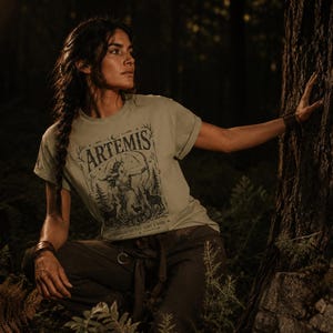 May include: Sage green t-shirt with the word "ARTEMIS" and a graphic of a woman with a bow and arrow, surrounded by trees and deer. The shirt is paired with dark green pants. The image is set in a forest with a dark, moody atmosphere.