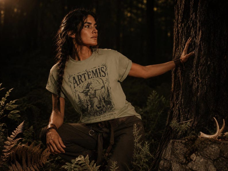 May include: Sage green t-shirt with the word "ARTEMIS" and a graphic of a woman with a bow and arrow, surrounded by trees and deer. The shirt is paired with dark green pants. The image is set in a forest with a dark, moody atmosphere.