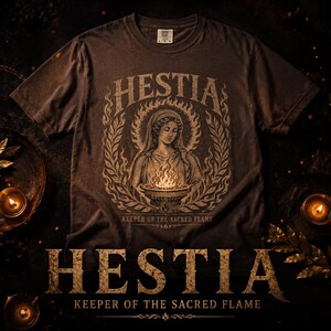 May include: Brown t-shirt featuring a detailed graphic of Hestia, the Greek goddess of the hearth, surrounded by a laurel wreath. The text "Hestia, Keeper of the Sacred Flame" is displayed in a vintage style.