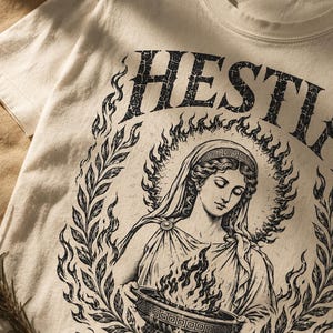 May include: Cream-colored t-shirt featuring a black graphic of a woman with a halo, surrounded by flames and laurel leaves. The word "HESTIA" arches above the figure. The design is vintage-inspired and printed on a soft, casual tee.