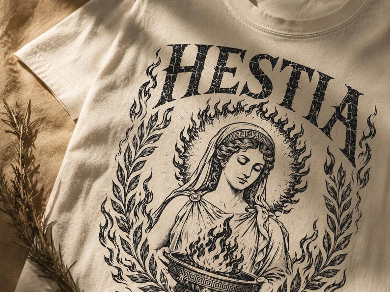 May include: Cream-colored t-shirt featuring a black graphic of a woman with a halo, surrounded by flames and laurel leaves. The word "HESTIA" arches above the figure. The design is vintage-inspired and printed on a soft, casual tee.