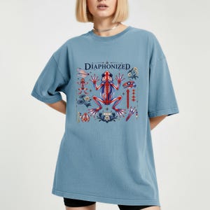 Könnte beinhalten: Ein staubblaues Kurzarm-T-Shirt mit einem zentralen Grafikdruck, der den Text "DIAPHONIZED" über einer detaillierten anatomischen Illustration eines Froschskeletts in Rot und Blau zeigt, umgeben von kleineren Skelettelementen und Blumenmotiven.