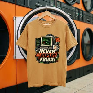 May include: A tan short-sleeve t-shirt featuring a graphic print of a vintage computer, a coffee mug, and the text "NEVER DEPLOY FRIDAY" in bold white and orange lettering, surrounded by various technical icons and coding symbols.