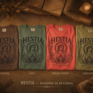 May include: Four crew neck t-shirts in espresso, sage, bright salmon, and light green. Each shirt features the word "HESTIA" above a graphic of a woman with a flame and the text "KEEPER OF THE SACRED FLAME". The text "HESTIA - Available in 14 Colors" is at the bottom.