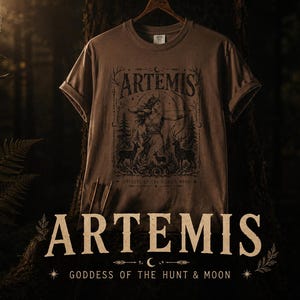 May include: A brown t-shirt featuring a detailed graphic of Artemis, the Greek goddess of the hunt and moon, with deer and forest elements. The word "ARTEMIS" is prominently displayed above the illustration, with "Goddess of the Hunt & Moon" below.