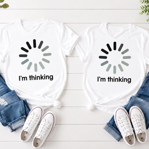 Peut inclure: Deux t-shirts blancs à manches courtes, chacun arborant un graphique circulaire de chargement noir et gris centré sur la poitrine au-dessus du texte noir "I'm thinking". Chaque t-shirt est associé à un jean bleu et des baskets blanches.