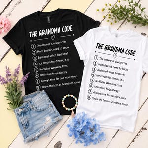 May include: A black t-shirt and a white t-shirt, each featuring the text THE GRANDMA CODE and a numbered list of eight rules including The answer is always Yes, Mom doesn't need to know, Bedtime? What Bedtime?, Ice cream for dinner, it is, No Rules Weekend Pass, Unlimited hugs always, Always time for one more story, and You're the boss at Grandmas house.