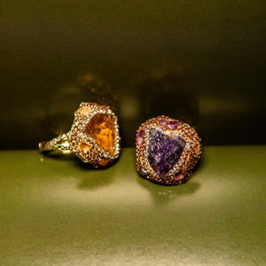 May include: Two rings featuring large, irregular raw gemstones. One ring displays a translucent amber-coloured stone, while the other showcases a deep purple amethyst-coloured stone. Both settings are encrusted with small, shimmering copper-toned crystals.