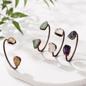 May include: Four open cuff bracelets made of twisted dark copper wire, each featuring two raw, uncut gemstone points at the ends. The stones include yellow citrine, pale green fluorite, translucent white quartz, and deep purple amethyst crystals.
