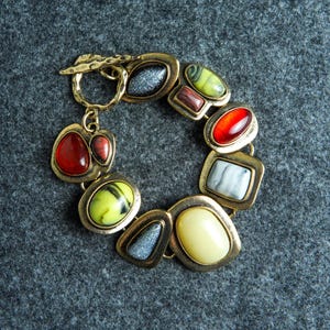 May include: A link bracelet featuring a series of multi-shaped, bezel-set cabochons in shades of red, yellow, green, white, and grey. The bracelet is constructed with a hammered gold-tone metal finish and secures with a toggle clasp closure.