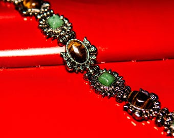 May include: A link bracelet featuring alternating square green stones and oval brown tiger eye stones set in a dark metallic frame. The design consists of repeating segments with intricate metalwork surrounding each gemstone, creating a continuous chain.