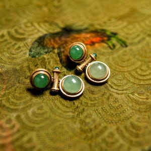 May include: A pair of gold-toned cufflinks featuring round, smooth, light green jade-like cabochon stones. Each cufflink consists of two circular settings connected by a central bar, with a small green accent bead on the post of each piece.