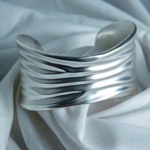 May include: A wide silver cuff bracelet featuring a matte finish and a series of horizontal, undulating ridges that create a textured, rippled appearance across the entire surface of the metal band, which has an open back design.