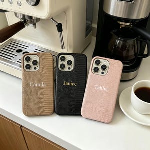 May include: Three smartphone cases with a textured faux-reptile skin pattern. The left case is tan with the name Camila, the center case is black with the name Janice, and the right case is light pink with the name Tahlia printed in gold lettering.