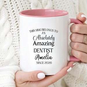 May include: A white ceramic mug with a pink interior and handle. The front features black text that reads: THIS MUG BELONGS TO AN Absolutely Amazing DENTIST Amelia SINCE 2026. The design uses a mix of bold sans-serif and elegant script fonts.