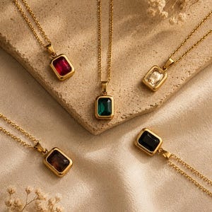 May include: Five gold-tone necklaces featuring rectangular pendants with faceted gemstones in red, clear, emerald green, dark brown, and black. Each pendant is suspended from a delicate gold-tone chain, showcasing a polished metal setting.