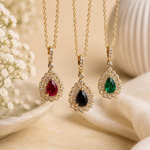 Può includere: Tre collane in oro con pendenti a forma di goccia. Ogni pendente presenta una pietra centrale rossa, nera o verde, circondata da un alone di piccole pietre chiare. Le catenine in oro sono sottili e le contromaglie sono decorate con pietre.