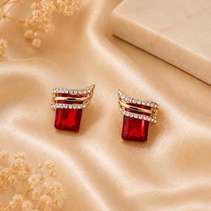 May include: A pair of gold-tone stud earrings featuring a rectangular red gemstone base. The upper portion of each earring is adorned with two curved, parallel gold bands encrusted with small, clear, sparkling white crystals.